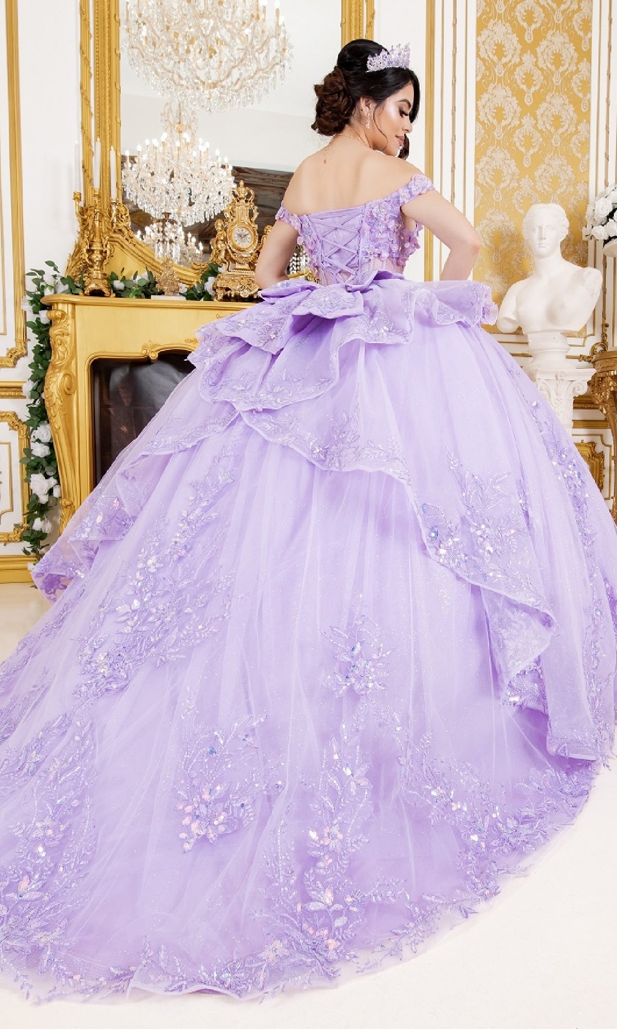 A woman in the Cinderella Couture 8160J off-the-shoulder lavender quinceanera dress with floral embellishments stands in an ornate gold room featuring a chandelier and marble statue.